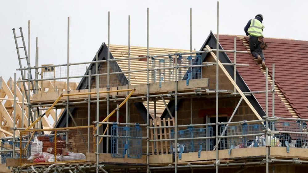 A man in a green high visibility jacket working on a roof of a house with lots of scaffolding round the structure. 