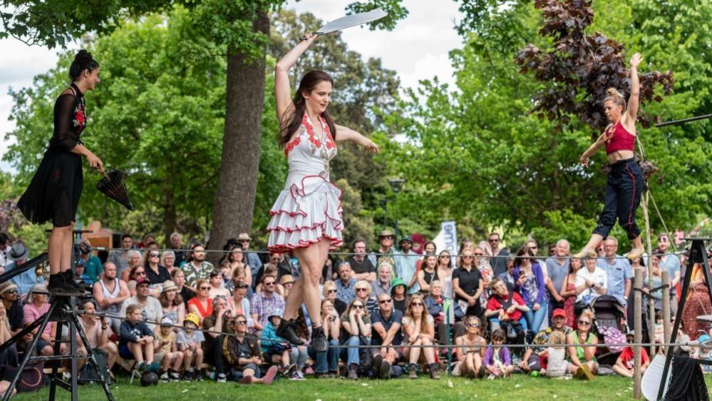 A troupe of three female wire walkers perform in front of a crowd in a park. The woman closest to the camera is holding a machete knife while balancing on the wire. 