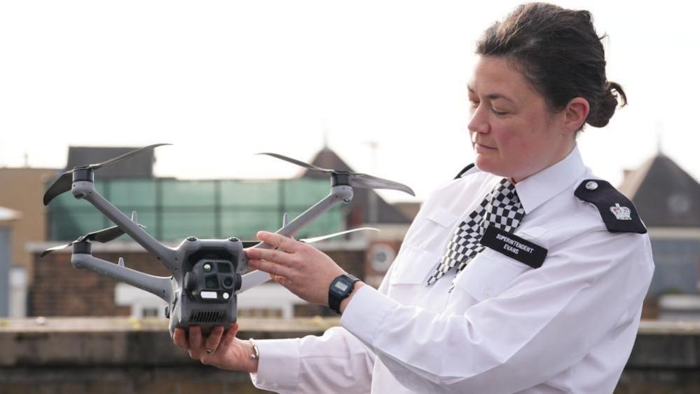A superintendent holds a grey drone while looking at it. She is wearing a white long sleeve shirt and a black and white chequered tie. Her hair is in a bun and she is wearing a black watch.