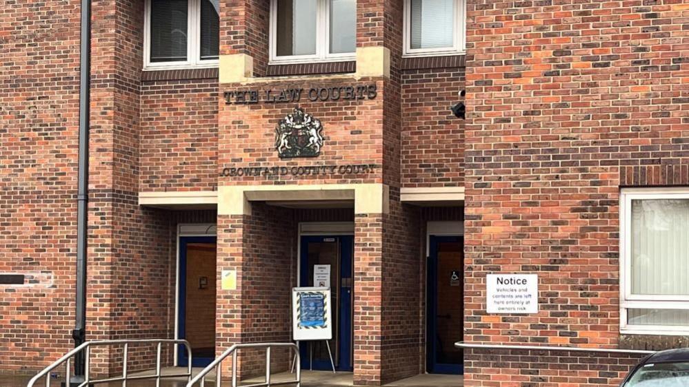 Norwich Crown Court main entrance, showing the red-brick building with stone lintels, a window, with some notices and the royal crest above the door. Chrome hand rails can be seen in the foreground.