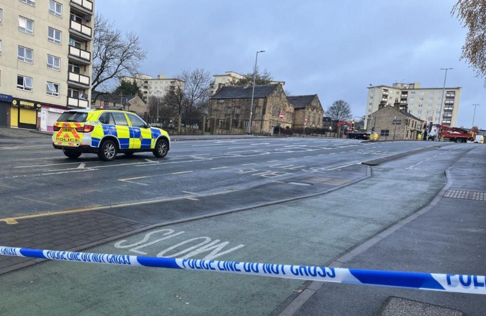 A police vehicle parked in the middle of a deserted street with blue and white police tape in the foreground.
