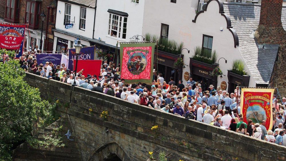 An aerial photo of people marching as part of a previous Miners' Gala. They're walking across a stone bridge with various colourful banners.