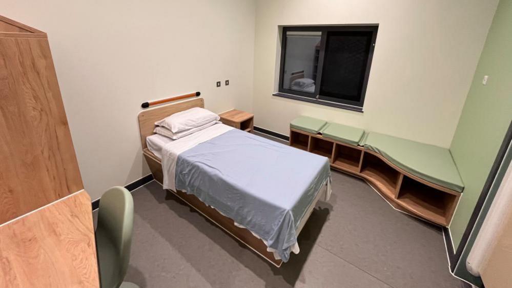 The interior of a mental health care suite inside the New Rivers Centre at Hellesdon Hospital, which is operated by the Norfolk and Suffolk Foundation NHS Trust. The bed has white sheets, and pillows, and a blue blanket. There is a bench, of wood effect, with green cushions. A chair and desk are visible on the right. A windows is in the centre on the back wall, black, owing to darkness outside.