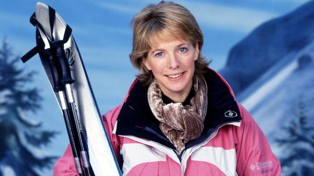 Hazel Irvine smiles as she holds skis in front of of an alpine background with snow-covered trees and the side of a mountain. She wears a pink ski jacket.