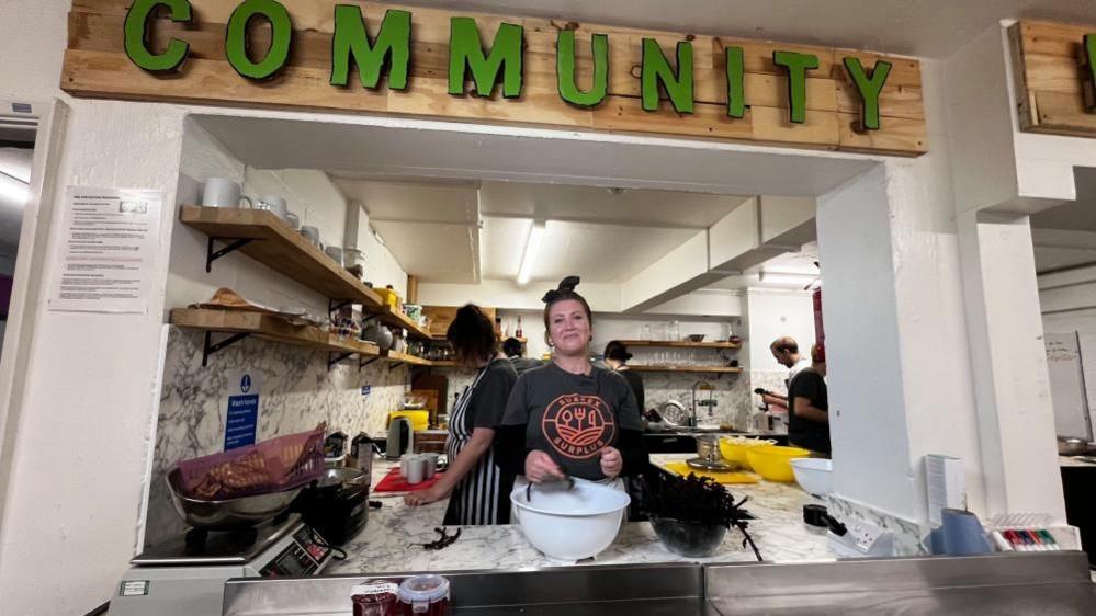 Ingrid Wakeling pictured in the Sussex Surplus kitchen. She is preparing food over a bowl. There are other chefs in the background working and a sign above her that reads 'Community'. 