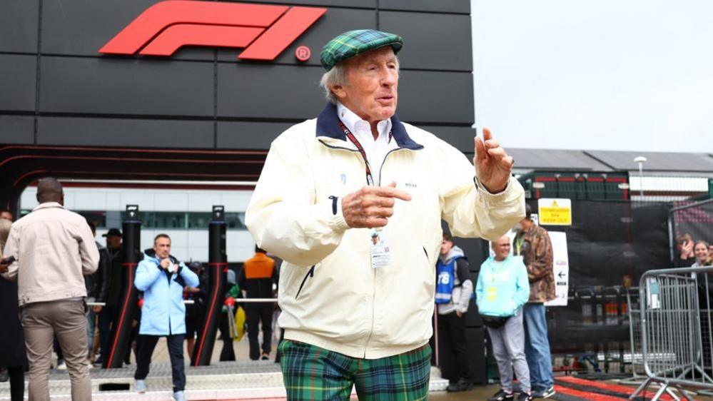 Sir Jackie Stewart in green tartan trousers, white jacket and green tartan flat cap. He is talking to someone off camera while in front of an F1 building.