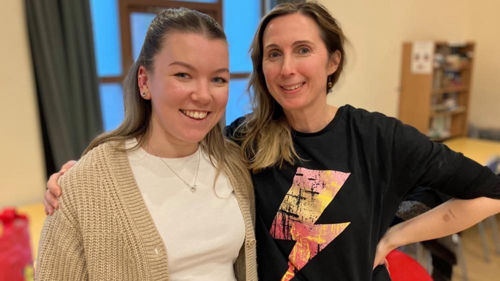 Two women stand and smile at the camera, in a village hall.