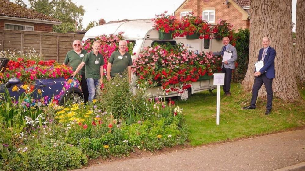 Members of Filby in Bloom stand together between an car and a caravan, which both form floral displays, with two judges, dressed in jackets with clip boards, standing closer to the tree. The area is bedecked with floral displays and lawns.