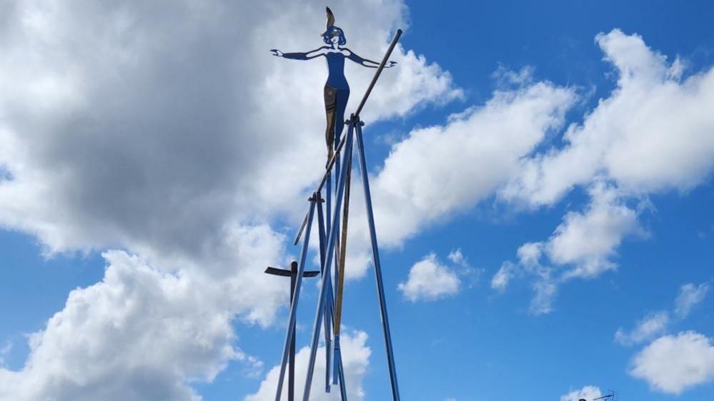 Tightrope, on Trafalgar Green, is a stainless steel trapeze frame with a flat sheet cut out of a woman, arms spread out and wearing a feather in her hair, balancing on a bar. The sculpture swings in the wind.