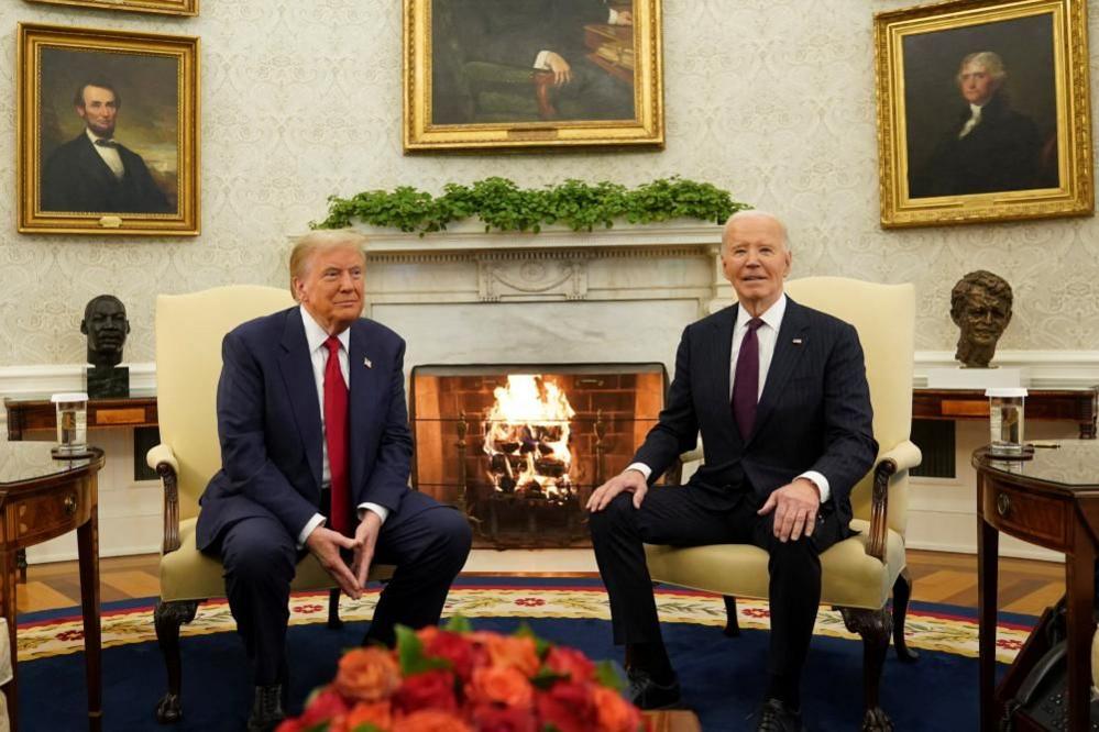 Donald Trump and Joe Biden seen sat on high-backed yellow chairs either side of a fire in the Oval Office. Portraits of past presidents are visible on the wall behind them. Both are looking out at reporters assembled out of sight behind the camera. 