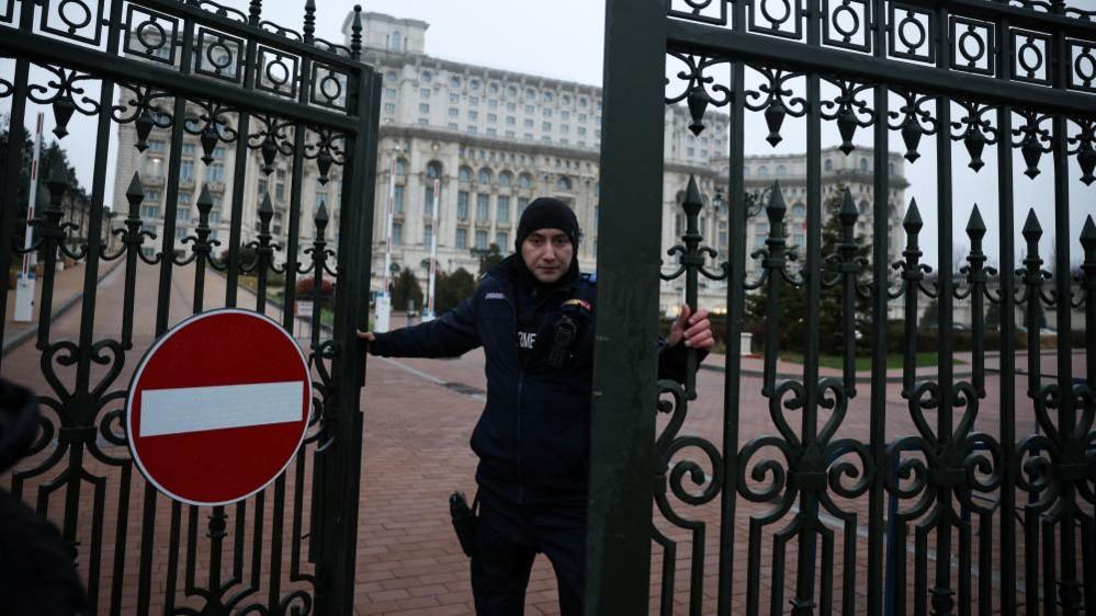 A police officer, wearing a dark hood and jacket, closes a wrought iron gate bearing a no entry sign at the grand Palace of Parliament