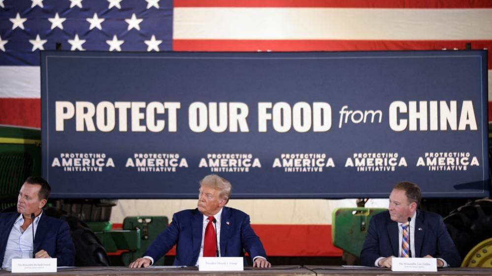 Donald Trump sits at a table with men in suits on either side and a large banner above his head which reads "Protect our food from China"