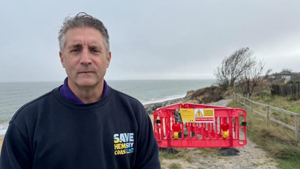 Simon Measures is wearing a purple polo shirt, the collar of which is visible under a blue sweat shirt embroidered with the Save Hemsby Coastline logo on his left breast. He is standing near the cliff edge in Hemsby at The Marrams, where orange fencing panels block a footpath.