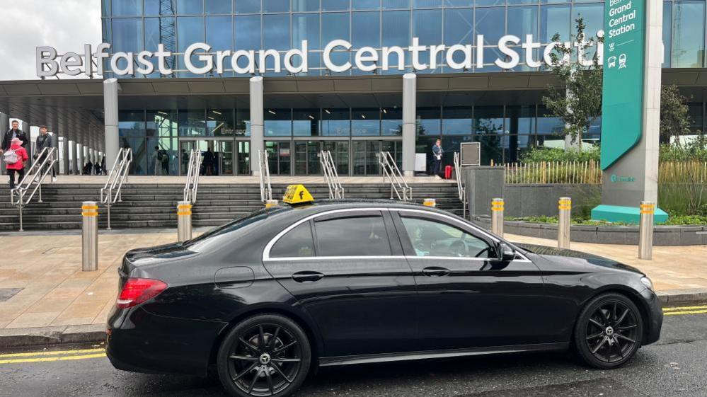A black car with a taxi sign on its roof is parked outside of the front entrance to Belfast Grand Central Station. The sign is white bold letters and there are steps up to a mostly glass building.