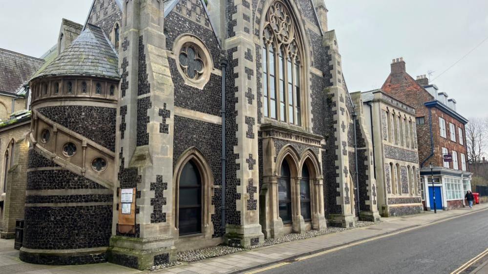 Greyfriars House is a former church and church hall, featuring a white sandstone and knapped flint facade. The building has a turret housing a staircase and large arched windows, and a chequerboard design at the upper parts of the gable ends.