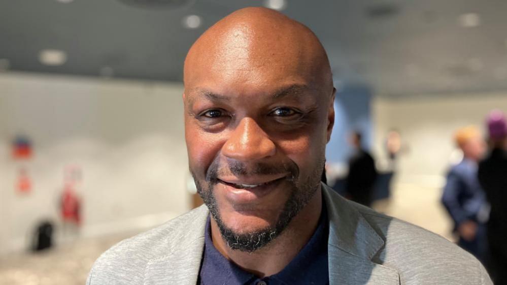 A face shot of Marcus Gayle, who is bald and has a short black beard. He is smiling at the camera, with a conference room in the blurred background.
