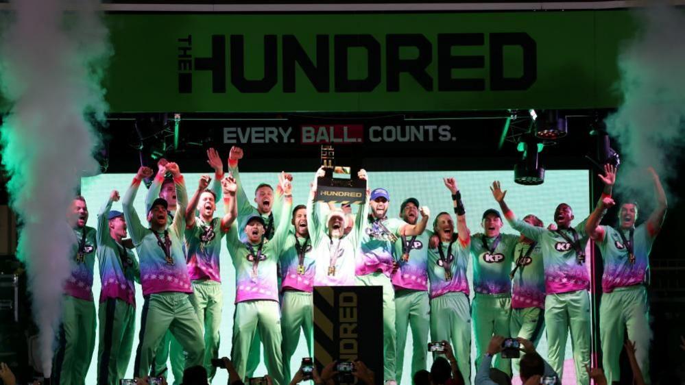 The Oval Invincibles cricket team lifting the Hundred trophy. Several cricketers in green celebrate on stage with smoke machines either side of them.