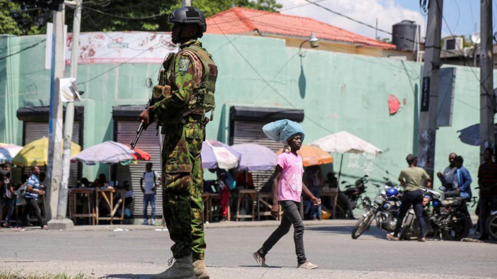 A Kenyan police officer can be seen on patrol. He is holding his weapon while a man carrying something on his head can be seen walking behind him. Other people are standing by their motorbikes.