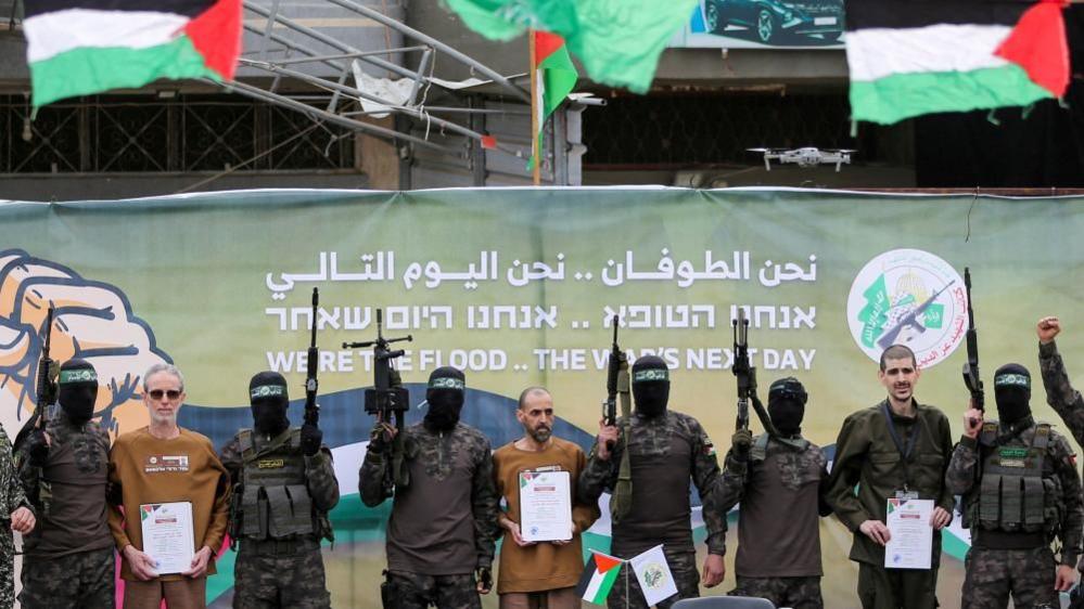 Armed and masked Hamas fighters pose with (L-R) Ohad Ben Ami, Eli Sharabi, Or Levy; the Hamas fighters hold their guns in the air and the Israeli hostages hold certificates. 