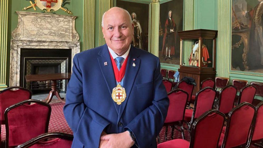 Lord Mayor of York Councillor Martin Rowley BEM stands in the main room at the Mansion House. He wears a blue suit, a blue tie and a ceremonial medal. In the background, rows of red seating can be seen, an ornamental fireplace, and large portraits hung on green walls.