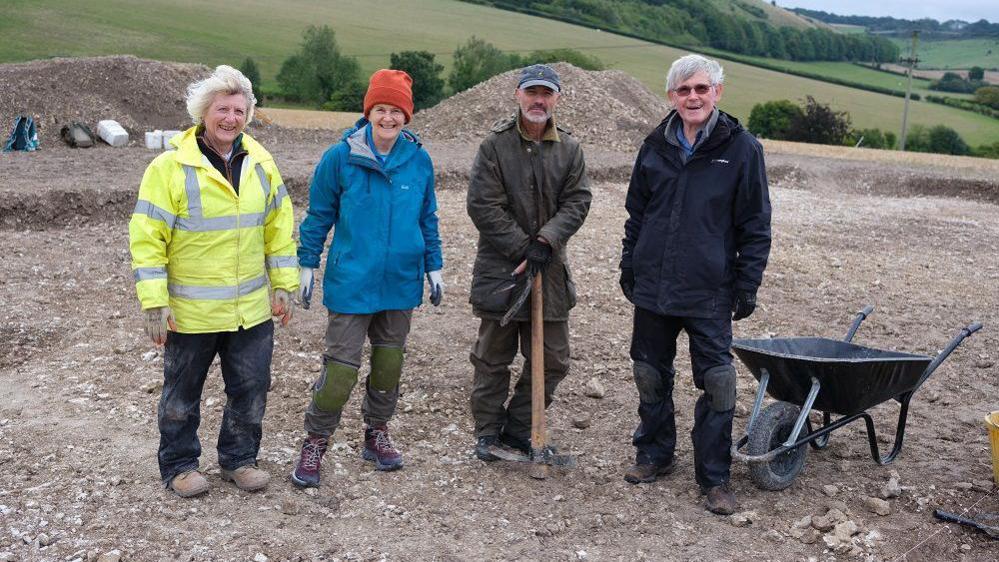 Four people standing in a large archaeology trench wearing large overcoats,  one with a pick-axe and a wheelbarrow to the side. Two large piles of soil behind them.