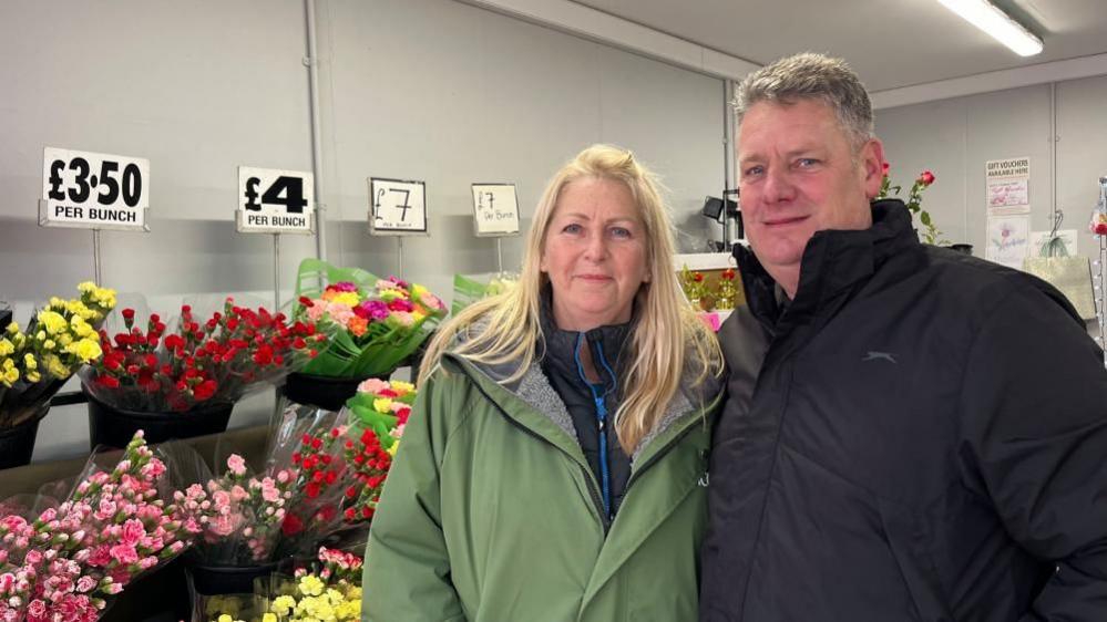 Sophie and Paul Howlett, standing beside each other inside their florist stall on Great Yarmouth Market. Beside them are bunches of flowers with prices displayed above. Sophie has long blonde hair, and is wearing a black jacket under a green coat with grey faux fur lining. Paul has greying brown hair, is clean shaven and is wearing a black jacket.