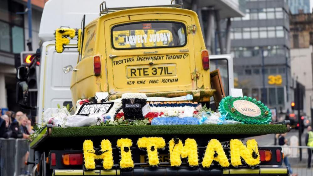 A yellow van is on the back of a flatbed truck driving down a road. A floral arrangement on the back of it spells 'HITMAN'.