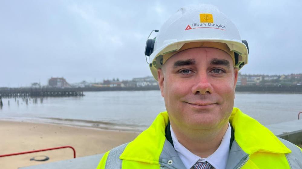 Councillor Daniel Candon, wearing a white hard hat and fluorescent yellow jacket, shirt and tie. He is smiling, standing near to the Spending Beach on the River Yare at Great Yarmouth's South Denes, with Gorleston Pier Hotel in the backdrop.