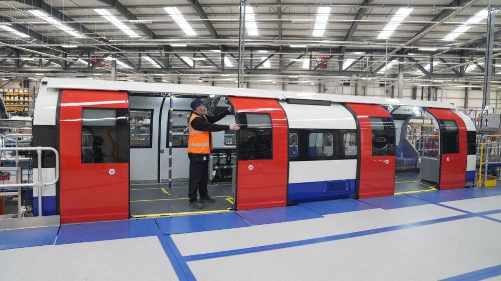 A blue, red and white train carriage inside a factory. The two sets of red doors of the carriage are open. A man wearing an orange hi-vis vest is stood in the doorway of the left set of doors, with his hands holding on to the side of one of the doors.