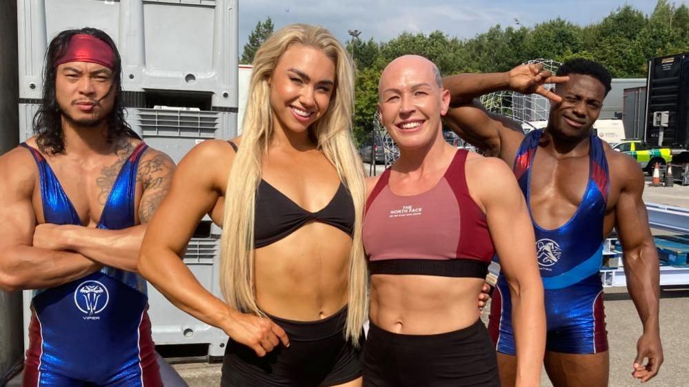 A female 'contestant' smiles joyfully, alongside three gladiators at an outdoor/ backstage location. She has a bald head, an athletic body and wears a crop top and shorts. The first male gladiator has long black hair and facial hair wears a shiny blue and red body suit and a red bandana. His arms are crossed and he has a menacing expression. The second male gladiator has short black hair and a muscular frame, and wears the same suit. He looks friendly and makes the 'V' peace sign with his hand.  The third gladiator is a tall female with long blond hair and a big smile. She has an athletic body and wears black shorts and a black bikini style top.