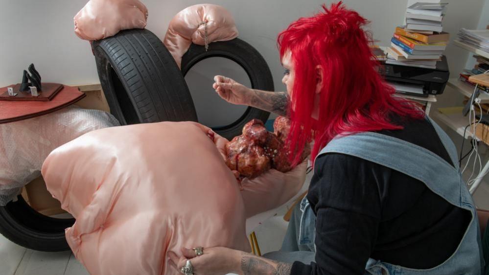 A woman with bright red hair, a black top and denim dungarees works on a peach coloured piece of art which involves a silk-like peach-coloured material and pearls. 