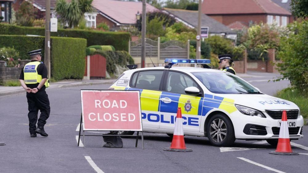 The image shows a police car parked diagonally on a road with houses along it. There is two traffic cones and a 'Road Closed' sign in front of the car. There is one police officer beside the car and one behind it.