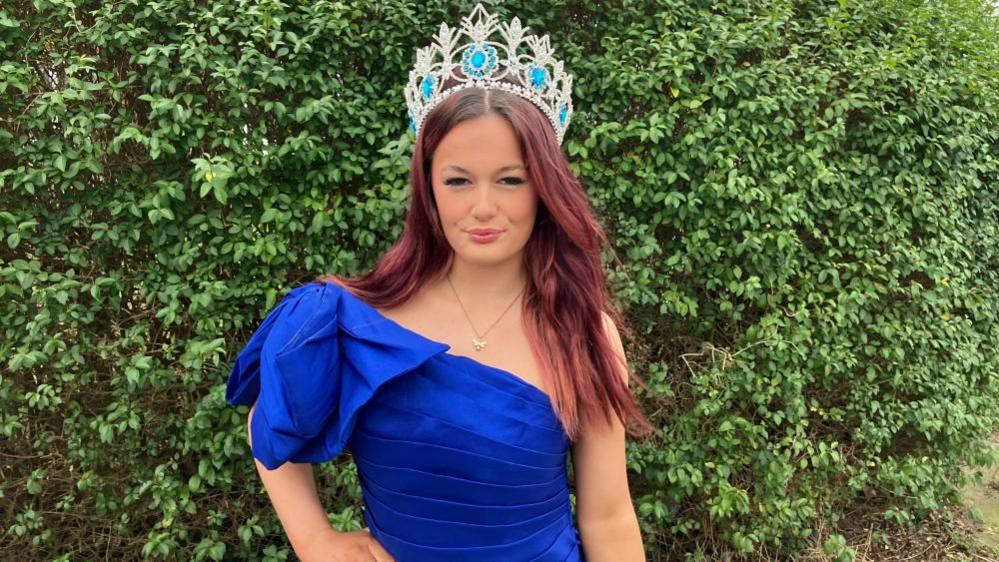 A teenage girl with dyed red hair stands outside wearing a blue ball gown and wears a large sparkly crown on her head. 