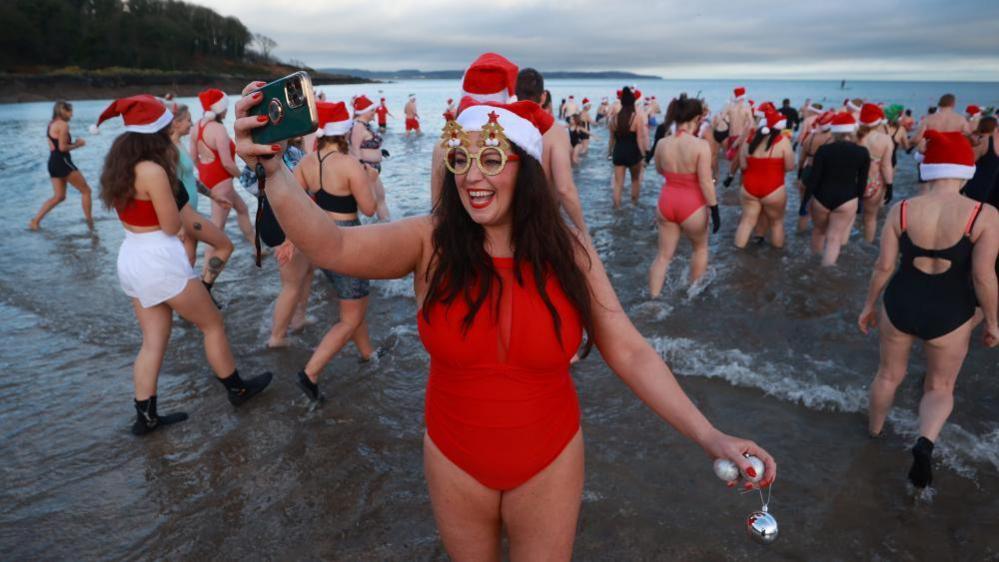 A woman wearing a red swimming costume is taking a selfie in front of a group of sea swimmers in the water.