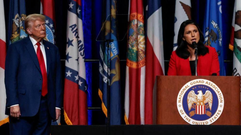 Tulsi Gabbard speaks at a lectern as Donald Trump watches on