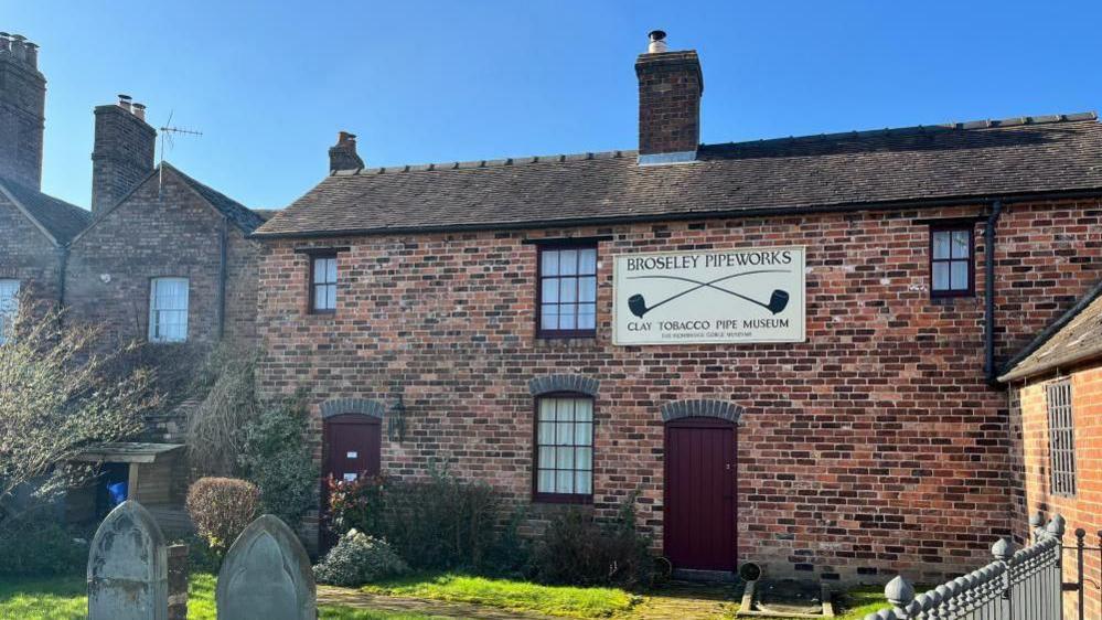 A brick building with two dark red doors and four windows - a large beige sign reads 'Broseley Pipeworks: Clay Tobacco Pipe Museum' in dark brown lettering. In the foreground is what looks like a small graveyard with bright green grass. The sky is bright blue and clear, with sunlight hitting the foreground of the image. 