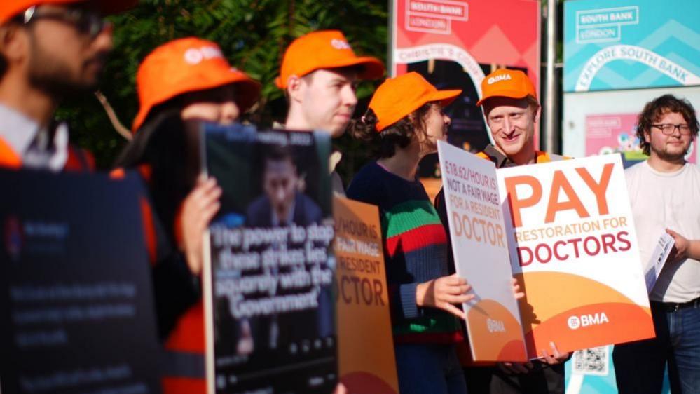 Doctors on a picket line. They are wearing orange hats and holding placards.