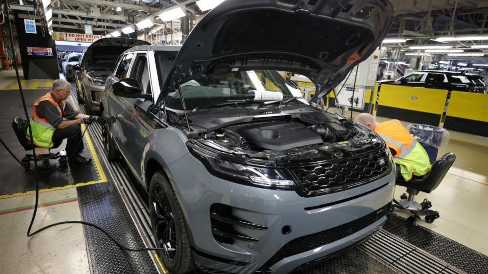 A grey land rover with its hood open sits on a factory floor, while two workers wearing hi-vis vests work on either side of the vehicle.