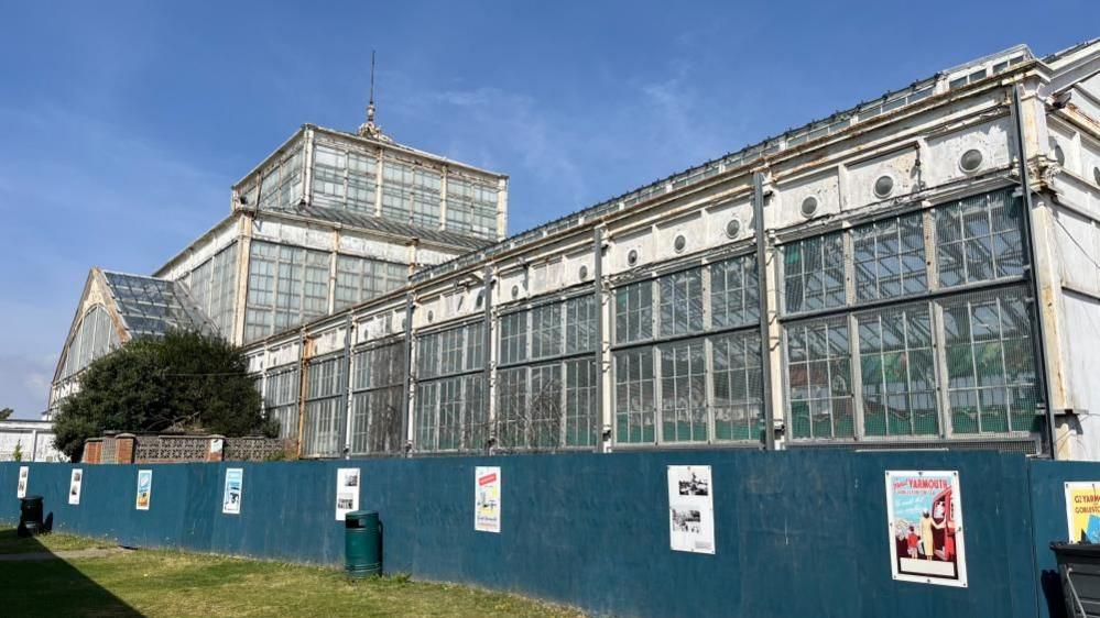 The Winter Gardens pictured from the south east, with the white ironwork and wood frames of the building, showing the lantern towards the centre and left of the image. There is a blue hoarding in the foreground with vintage posters promoting Great Yarmouth through the ages, displayed along it.