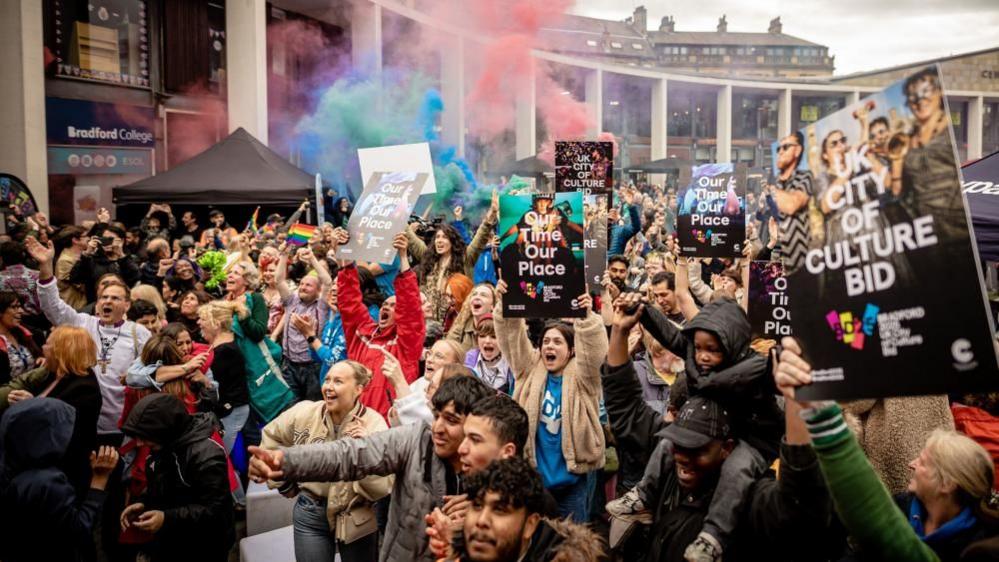 More than 100 people celebrate the news that Bradford would be City of Culture in City Park. They are holding placards that read: Our Time Our Place and UK City of Culture Bid.