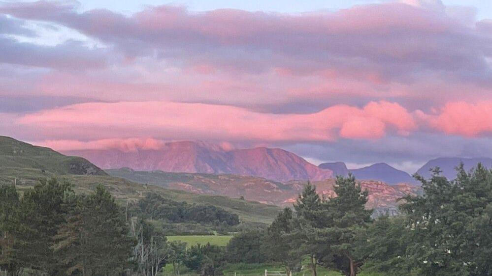 Large pink clouds hang over mountains in the distance. Closer to the viewer are green fields and hills and trees.