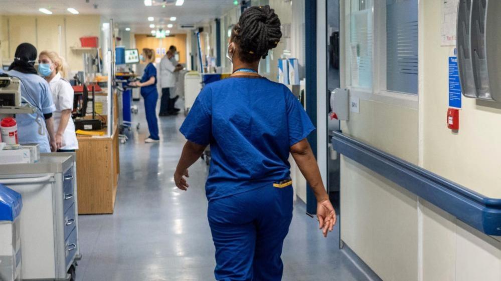 A nurse in blue scrubs walks through a busy hospital corridor