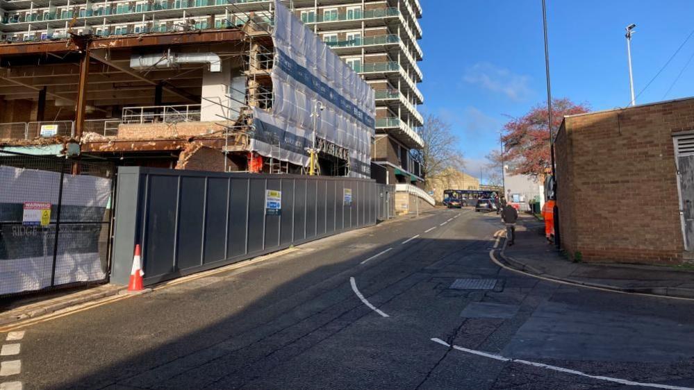 Wellington Street, Northampton. A road, showing a building being demolished to the right, a brick wall to the left and a man walking up the street. There are two lanes of traffic.