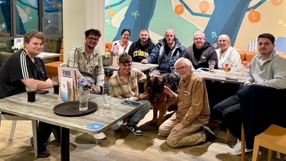 A group of people in a caravan park lounge, sitting on sofas and chairs, with a dog in the centre.