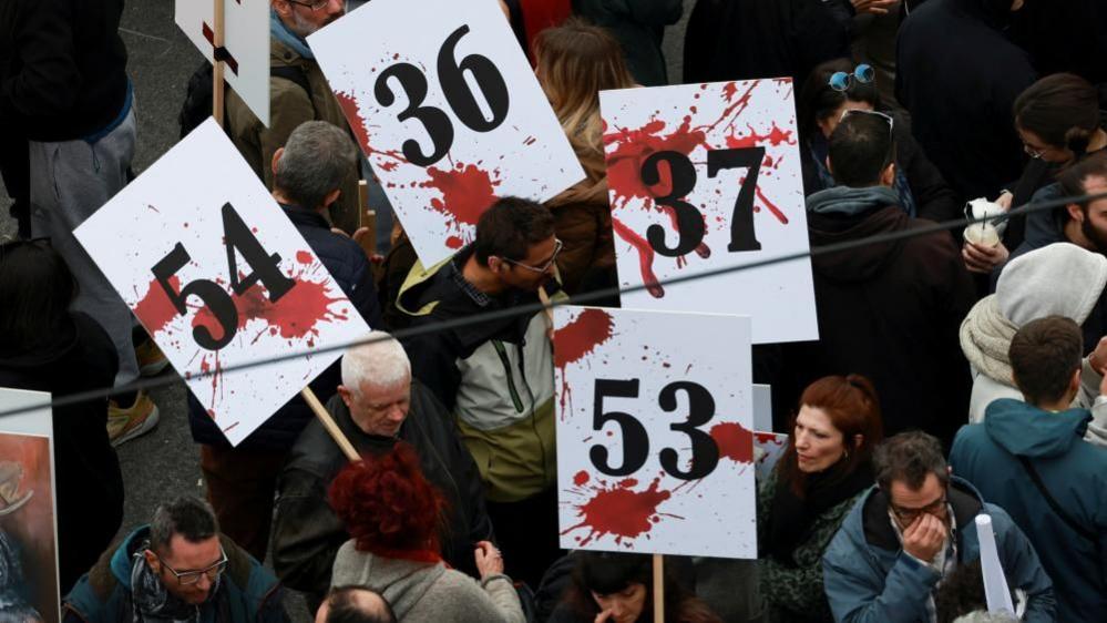 People holding white placards with red splodges and numbes 54, 53 and 37 to remember the dead