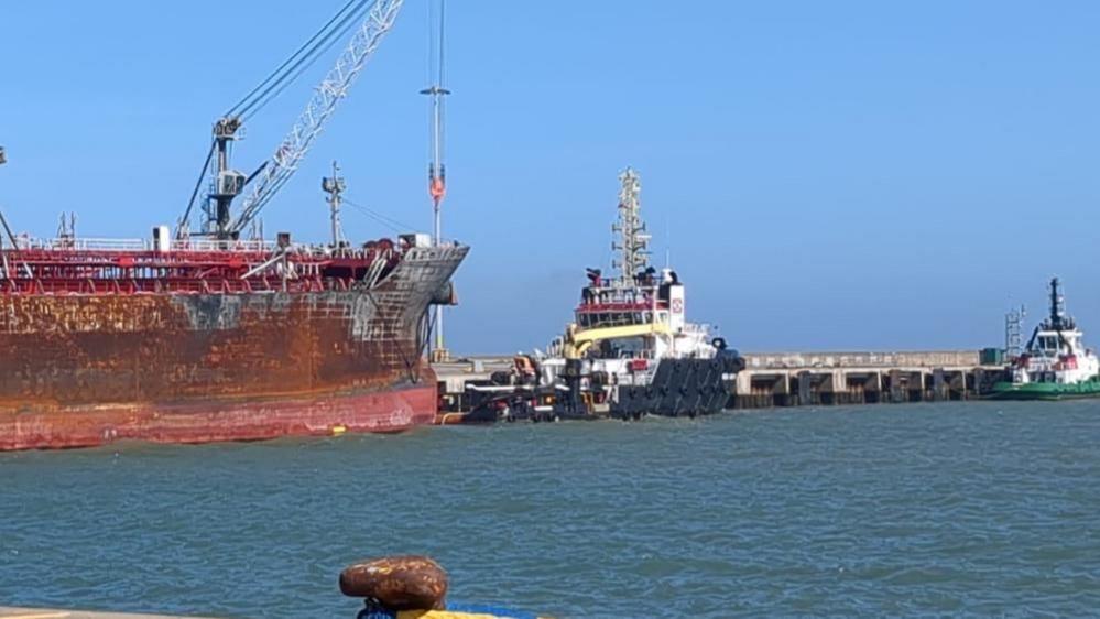 The Stena Immaculate rests on the North terminal of Great Yarmouth's outer harbour, with a tug at the bow. The hull is rusting and fire damaged following a collision in March off the Humber Estuary. Another tug is further towards the right of the image with quayside hardstanding in the foreground.