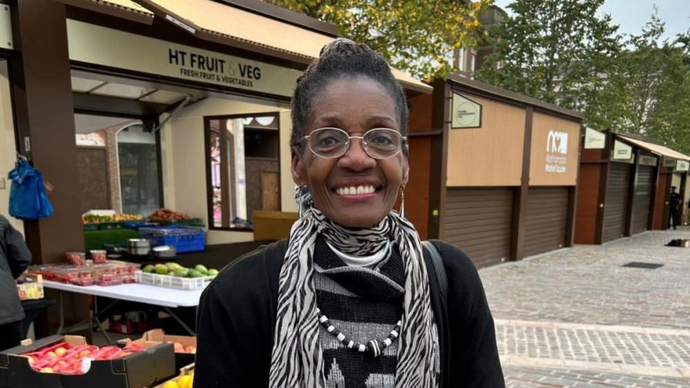 Morcea Walker with black hair and glasses, smiling and wearing a black top and black and white scarf with a fruit and veg stall in the background