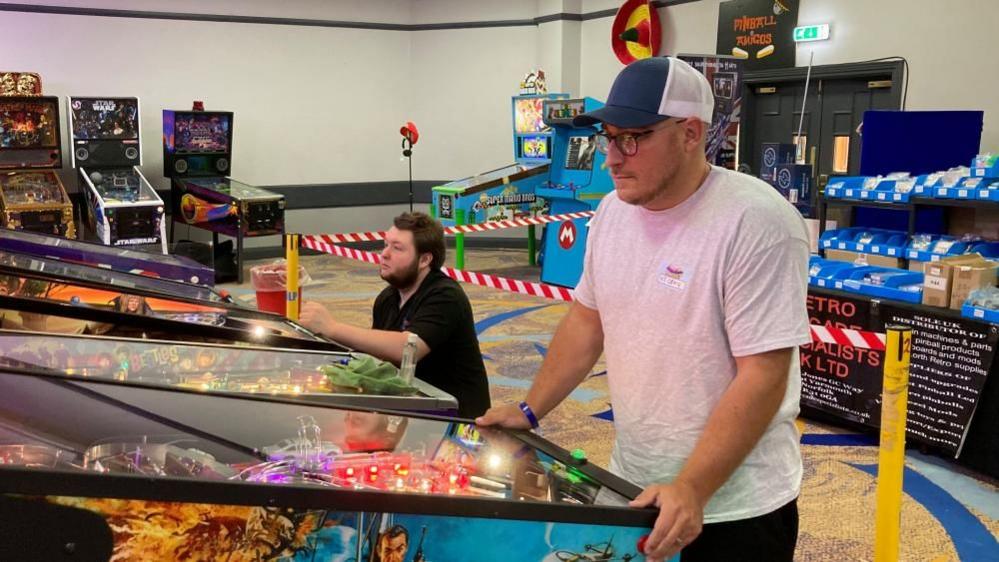 Craig Pullen wearing a blue and white cap standing by a mainly blue pinball machine