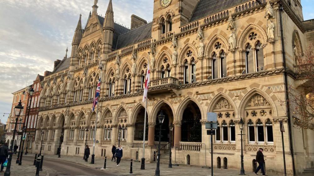A gothic Victorian building, showing a light stone building, with many small windows, a number of people walking past, two flags outside the building, black bollards and signs. There is a large entrance, with steps leading up and three arches, without windows in. 