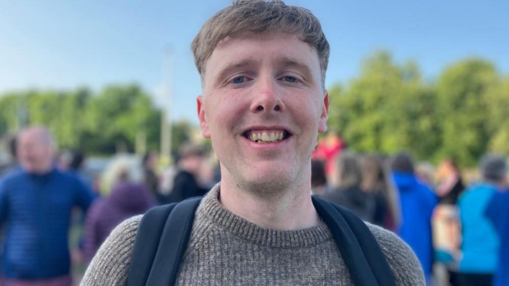 A man smiles into the camera. He has fair hair and is wearing a grey jumper. He has black backpack straps over his shoulders.